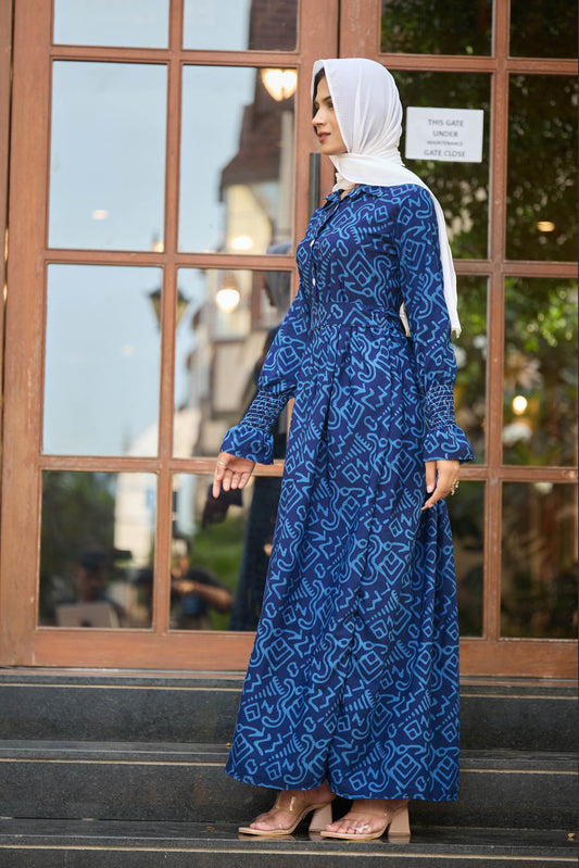 Woman wearing a blue patterned dress and white hijab standing in front of a glass door.